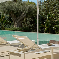 Light beige lounge chair with a sleek design, set against lush greenery and a tranquil aqua pool background.