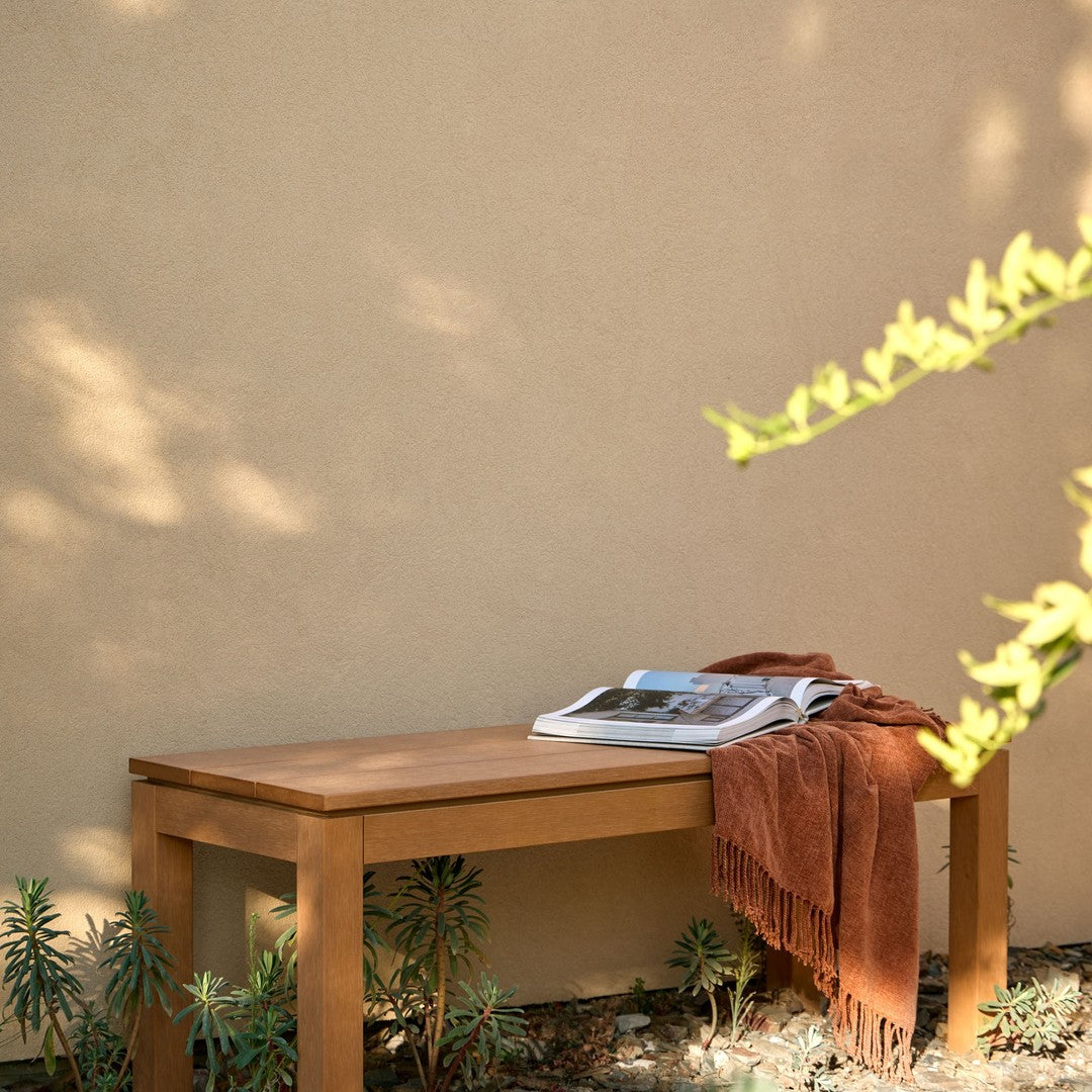 Natural wood bench with rich brown tones, paired with a rust-colored throw and greenery against a neutral wall.
