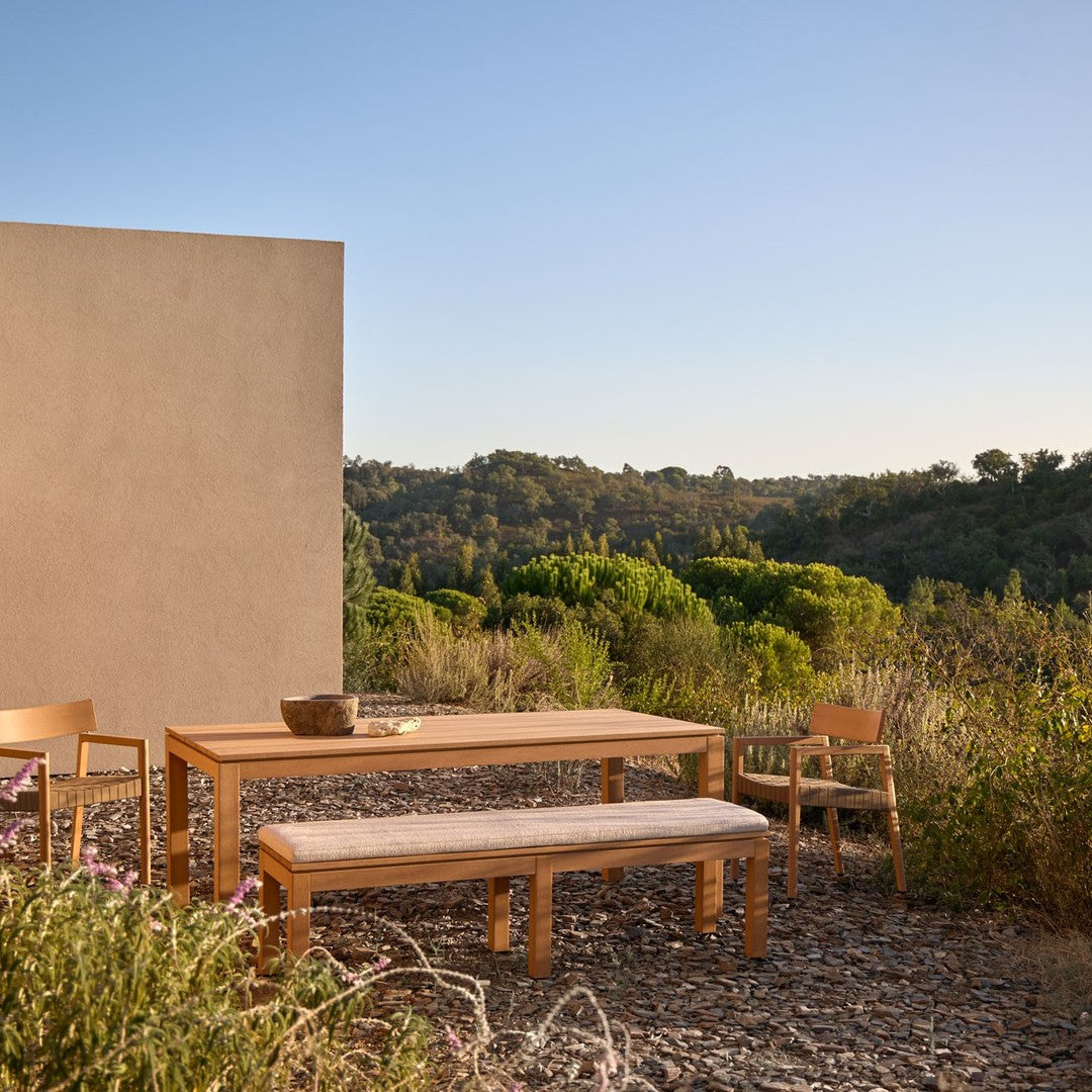 Natural wood dining set with light beige cushions, set against a serene green landscape and clear blue sky.