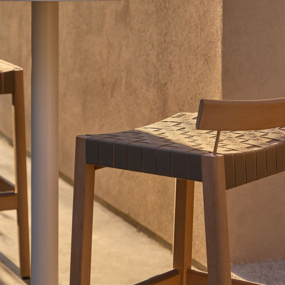 Brown woven bar stool with a geometric pattern on the seat, set against a textured beige wall.