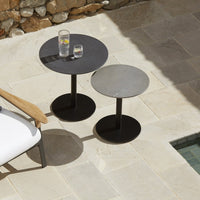 "Two round metal side tables in black and gray, set on a stone patio with light beige tiles."