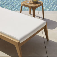 White cushion on a light wood frame, paired with a wooden side table beside a shimmering blue pool.