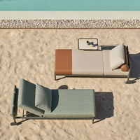 "Two modern lounge chairs in teal and beige with a brown accent, set on a sandy surface beside a pool."