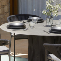 Modern round dining table with grey surface, black tableware, and textured black chairs, set against a natural stone backdrop.