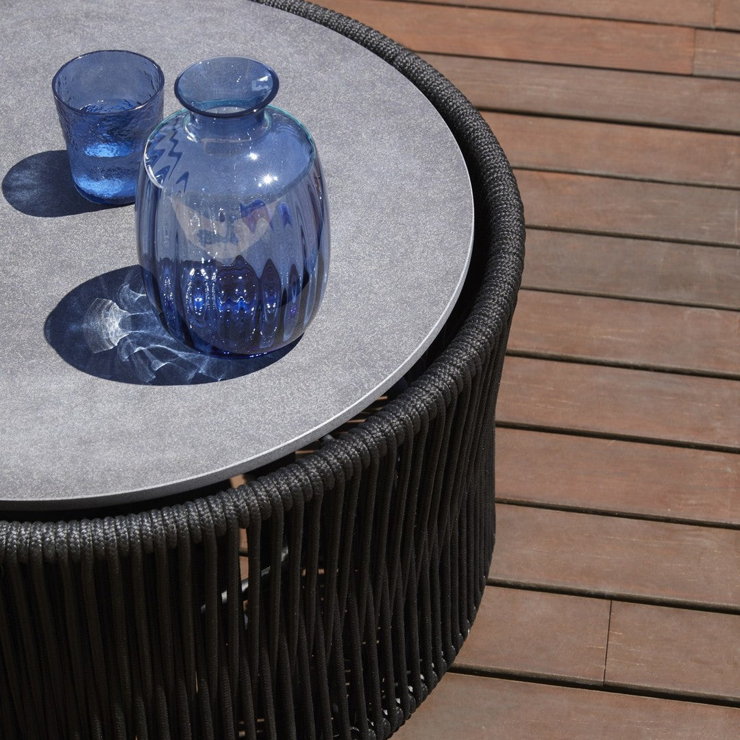Round black wicker table with a light gray top, featuring blue glass vases and glasses, set against wooden deck.