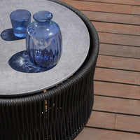 Round black wicker table with a light gray top, featuring blue glass vases and glasses, set against wooden deck.