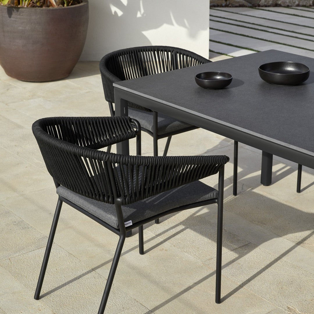 Black woven chairs paired with a sleek black table, set on a beige stone patio. Modern outdoor furniture design.