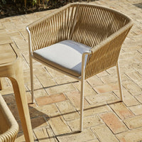 Tan woven chair with white cushion, featuring a modern, minimalist design on a rustic brick patio.