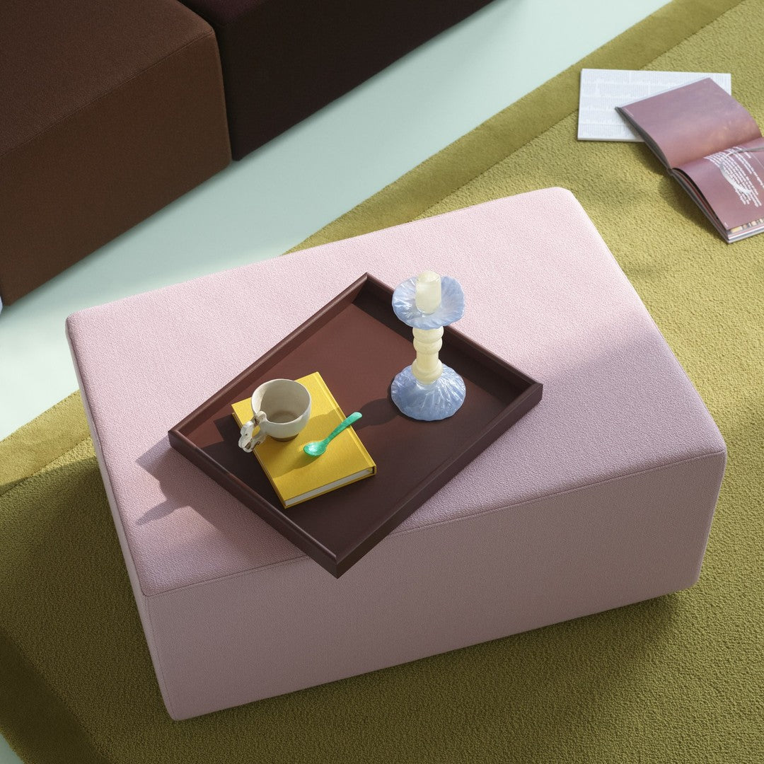 Pink square ottoman on green rug with dark brown accents. Tray features yellow and brown items, plus a white candle holder.