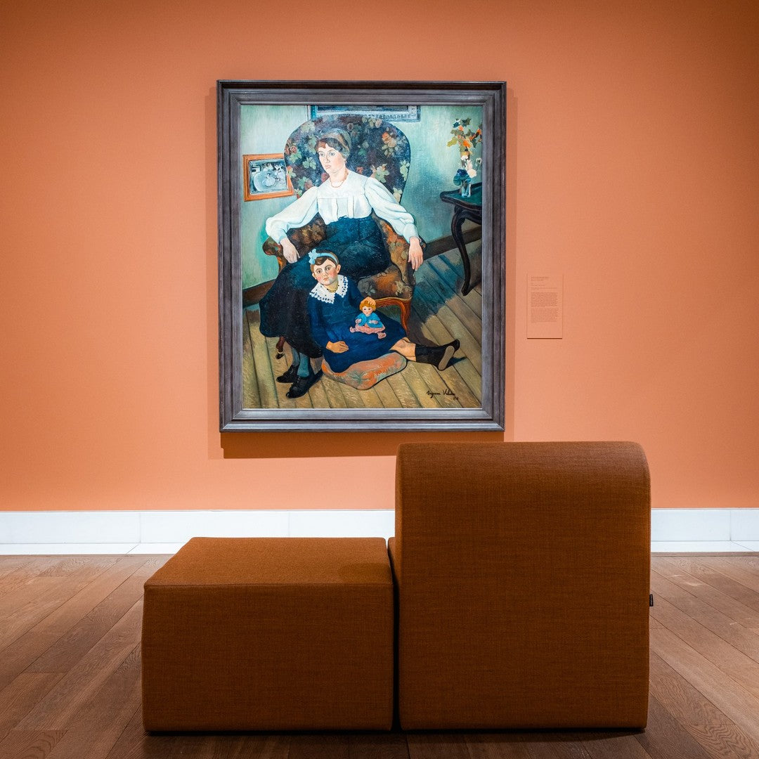 Casual brown fabric chair in front of an orange wall, with a colorful painting featuring vibrant blues and earth tones.