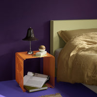 Orange metal side table with geometric design, set against a deep purple wall and a neutral beige bedding backdrop.