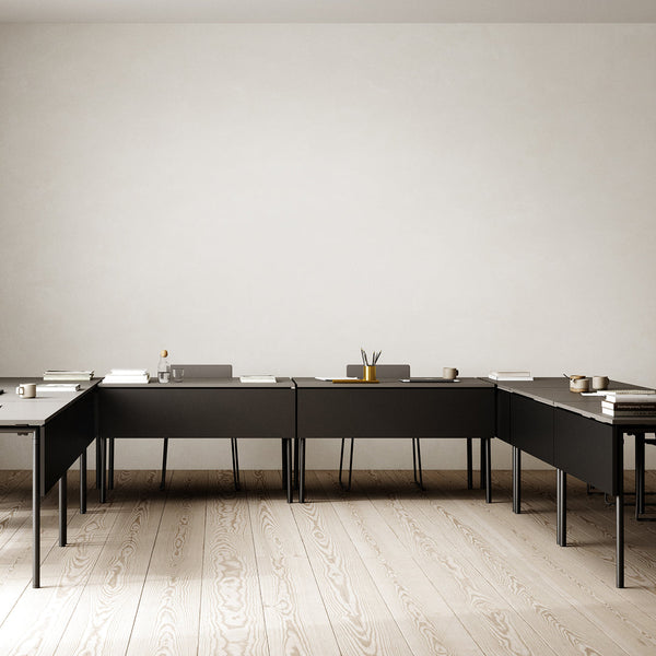 Modern black desks arranged in a minimalist workspace, featuring a light wood floor and neutral wall.