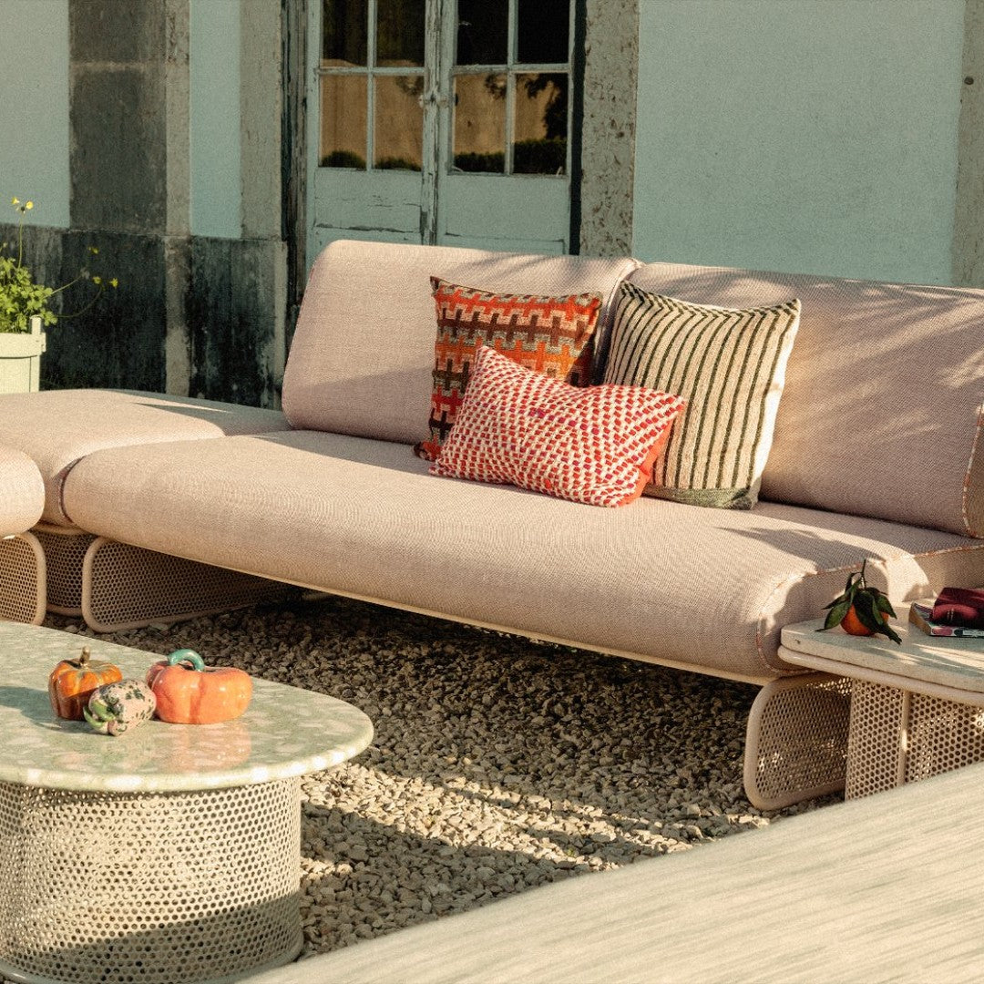 Pink sofa with patterned cushions in red, orange, and black; circular glass table and pebbled ground.