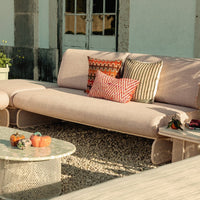 Pink sofa with patterned cushions in red, orange, and black; circular glass table and pebbled ground.