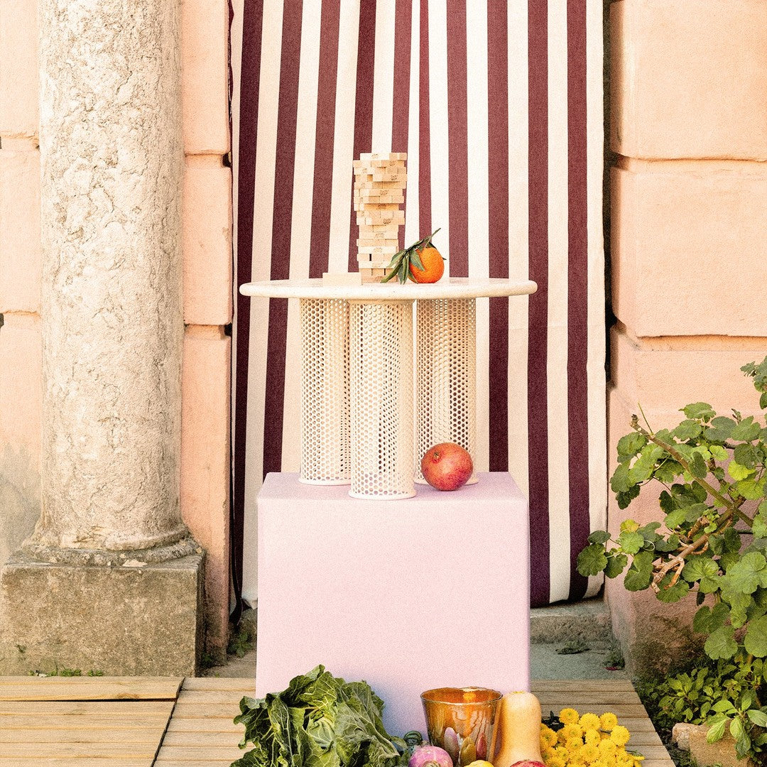 "Round wooden table with a cream top and white mesh legs on a pastel pink pedestal, against a striped burgundy backdrop."