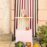 "Light wood table with mesh accents, set against a backdrop of burgundy and cream striped fabric, surrounded by colorful produce."