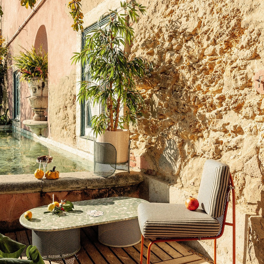 Gray striped chair with a red frame next to a round, green patterned table in a sunlit outdoor setting.