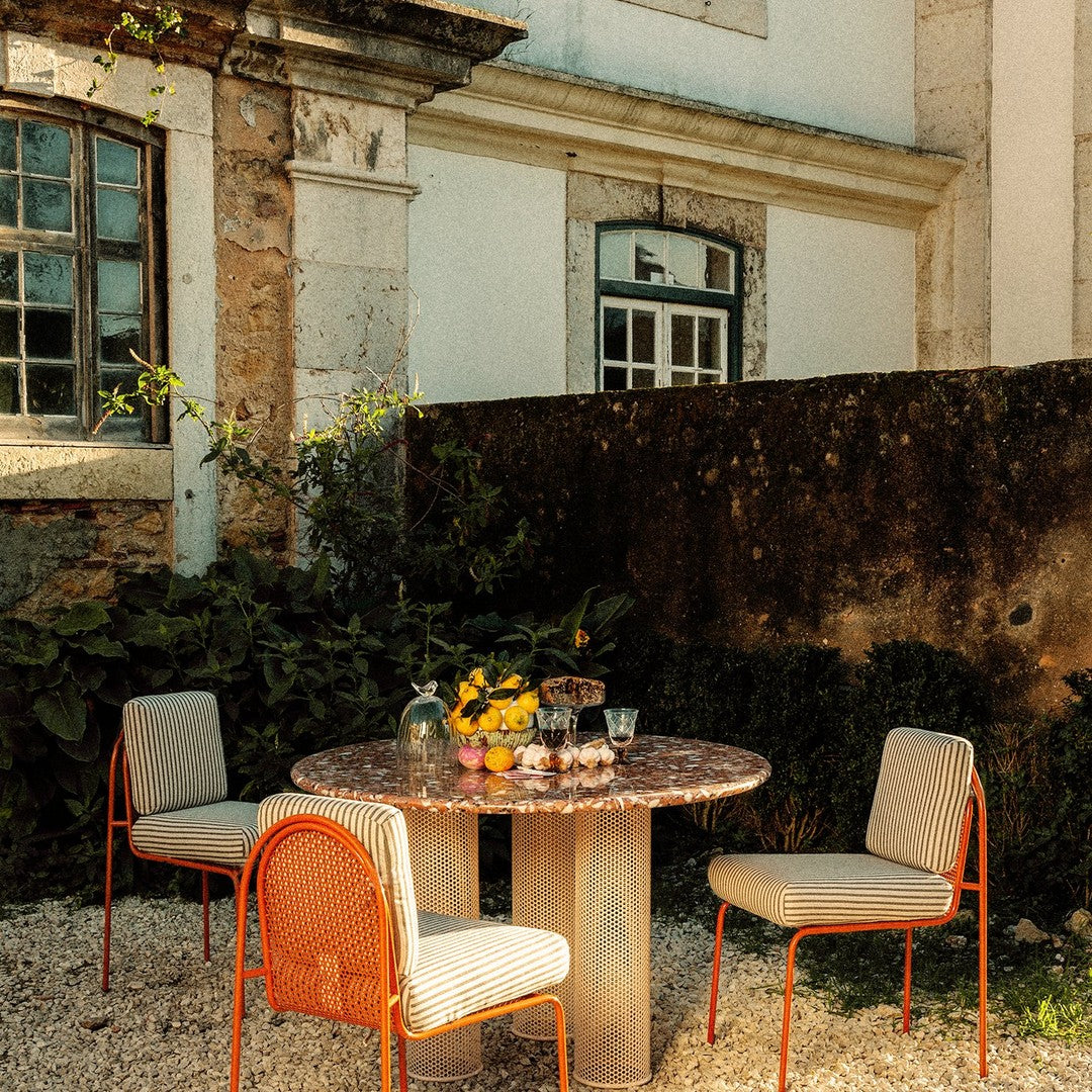 "Round table with a stone top, surrounded by beige chairs with orange accents, set in a rustic outdoor garden."