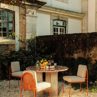 "Round table with a stone top, surrounded by beige chairs with orange accents, set in a rustic outdoor garden."