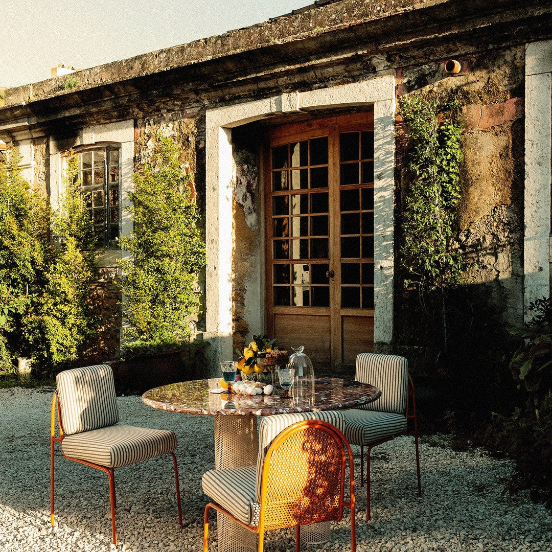 Outdoor dining set featuring a round stone table, patterned chairs in soft pastels, against a lush green backdrop.