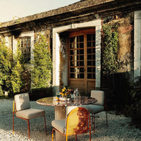 Outdoor dining set featuring a round stone table, patterned chairs in soft pastels, against a lush green backdrop.