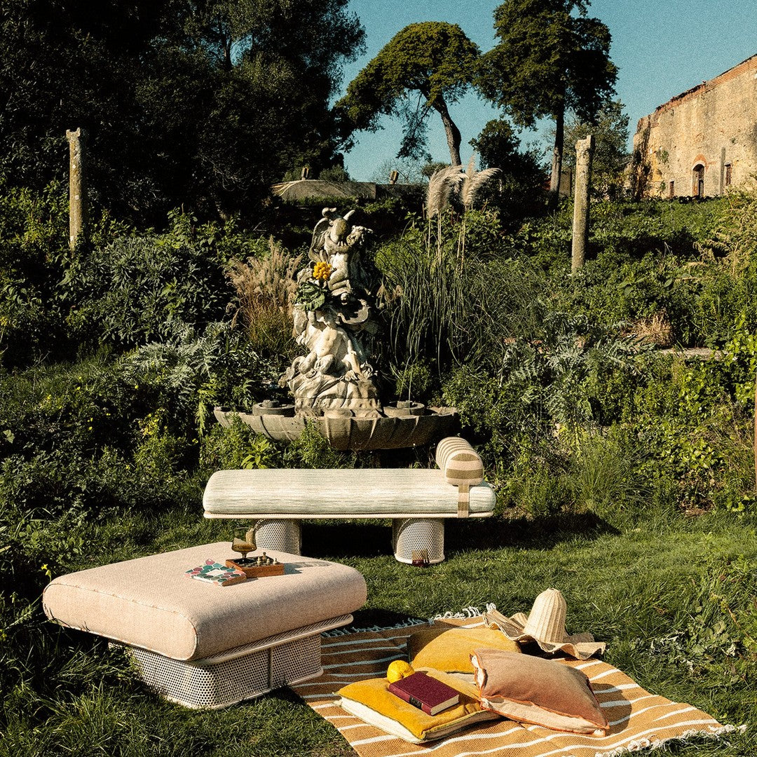 "Soft beige and yellow cushions with a striped blanket, set against lush greenery and a stone fountain backdrop."