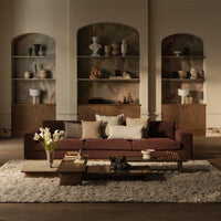 Brown velvet sofa with cream and beige cushions, set against a minimalist backdrop of warm tones and textured patterns.