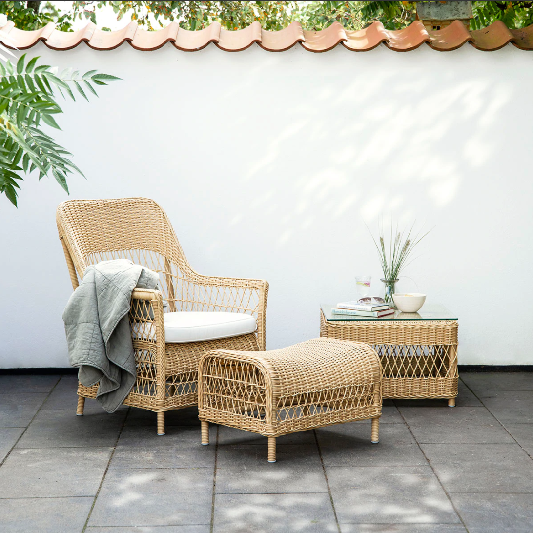 Natural woven rattan chair and ottoman set with a light beige cushion, accented by green foliage and a neutral backdrop.