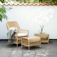 Natural woven rattan chair and ottoman set with a light beige cushion, accented by green foliage and a neutral backdrop.