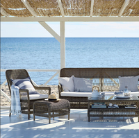 Coastal outdoor furniture set featuring rich brown wicker with soft white and blue cushions against a serene sea backdrop.