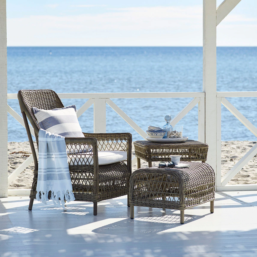 Woven brown wicker chair and table with a blue and white striped blanket and cushion by the beach.