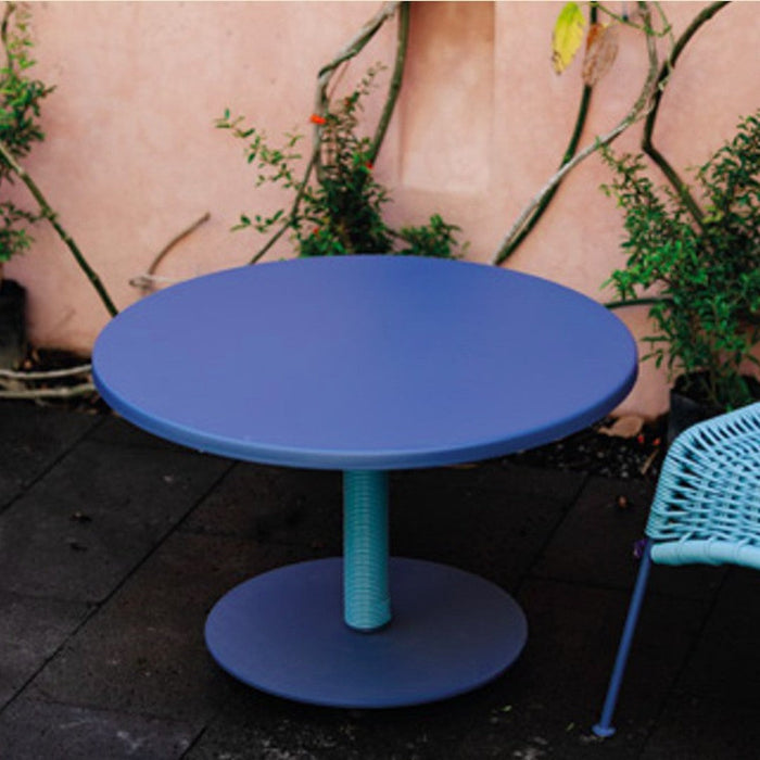Round blue outdoor table with a smooth top and a turquoise pedestal, set against a soft pink wall.