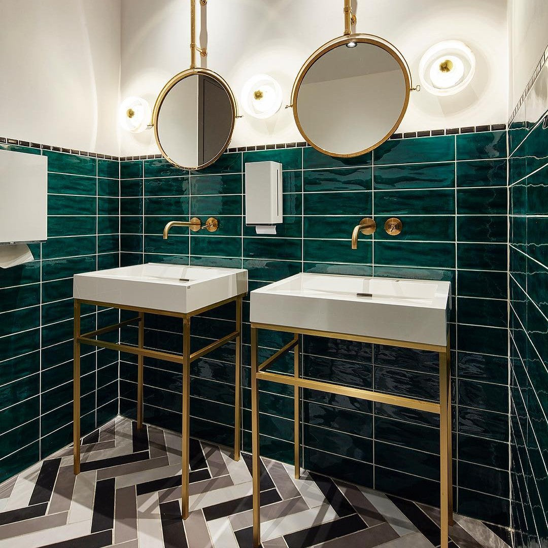 Chic green tiled bathroom with white sinks, gold fixtures, and a bold black and white herringbone floor pattern.