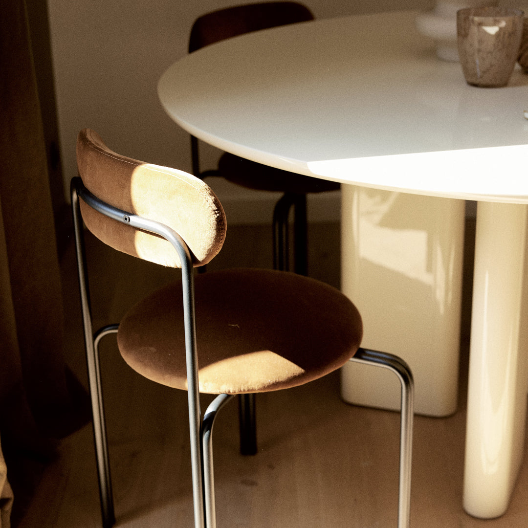 Brown velvet chair with a circular seat and backrest, metal frame, next to a white table in a warm, sunlit setting.