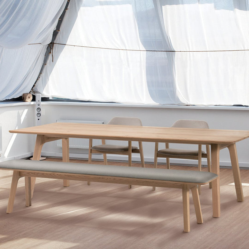 Light wood dining table with beige seating and soft fabric bench, set against a bright, airy backdrop.