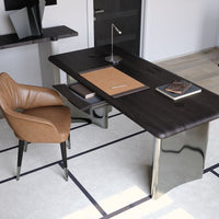 Modern black wood desk with sleek chrome legs, paired with a warm brown upholstered chair on a light beige rug.