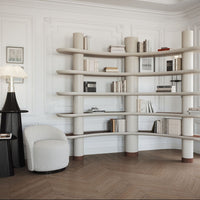 Curved beige bookshelf with smooth surfaces, accented by light wood and a neutral-toned armchair in a stylish room.