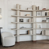 Curved beige bookshelf with light wood shelves and a textured white armchair against a neutral background.