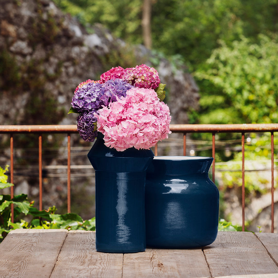 Navy vases with vibrant pink, purple, and blue hydrangeas on a wooden table. Lush green background.