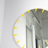 Circular mirror with vibrant yellow sunburst pattern, reflecting light against a modern white wall.
