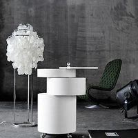 White circular side table with sleek layers, accompanied by a translucent white lamp and a green quilted chair.