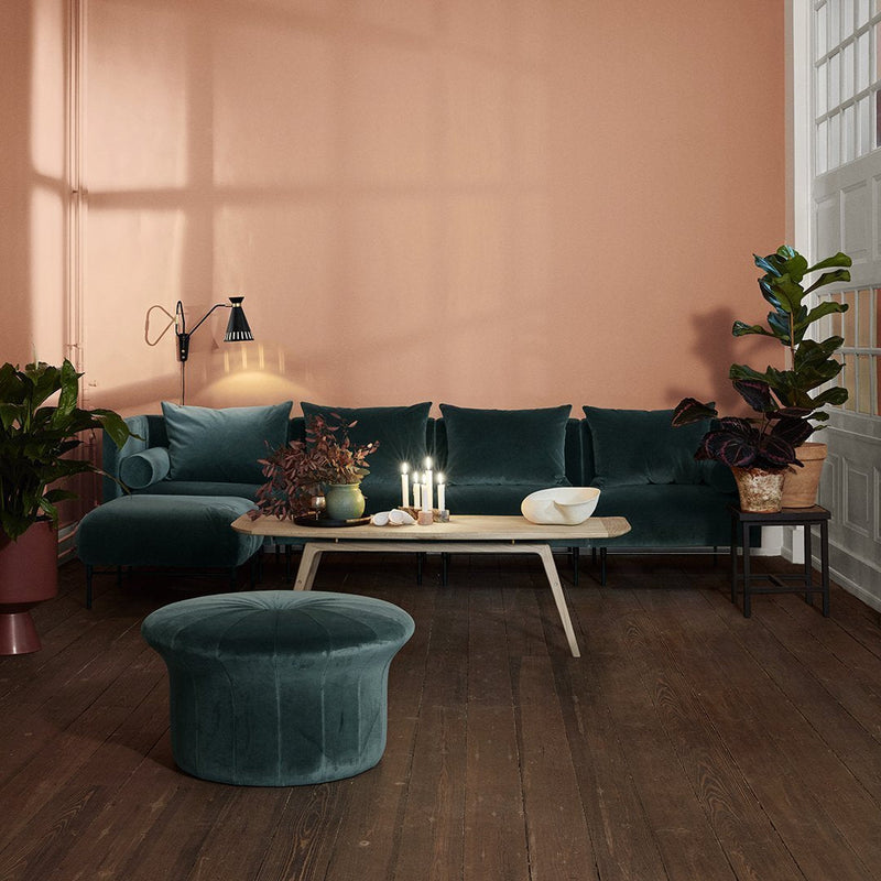 Teal velvet sectional sofa with round ottoman, set against a coral wall, complemented by green plants and wooden accents.